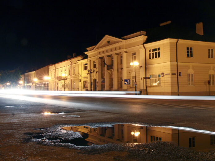 Kałuża na ziemi, odbijająca światło, w tle budynek nocą.