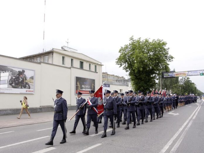 Funkcjonariusze zakładu karnego, w mundurach, idą środkiem ulicy trzymając sztandar