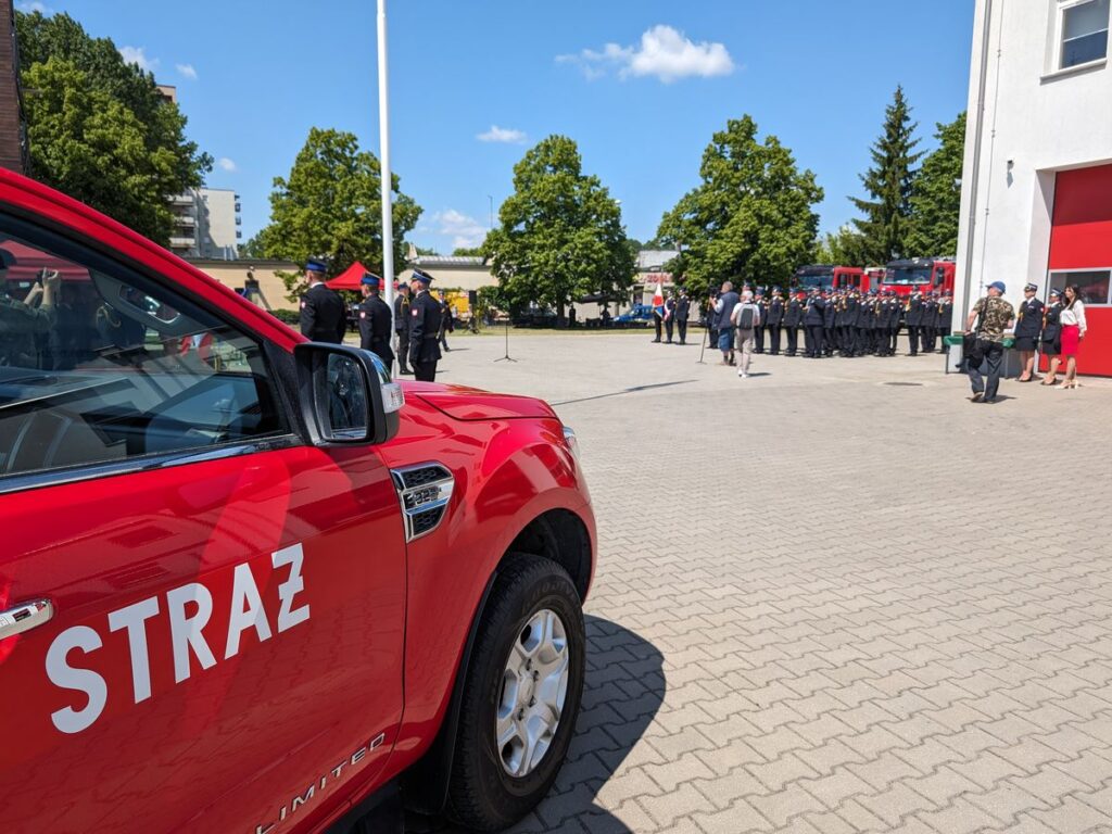 Czerwony samochód strażacki na pierwszym planie, a w tle zastępy strażaków