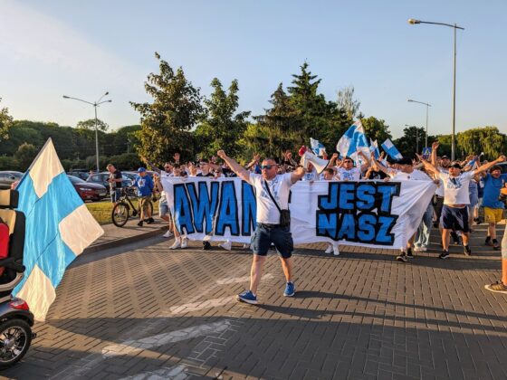 Młody mężczyzna z rozłożonymi rękami idzie przed transparentem z napisem "awans jest nasz"