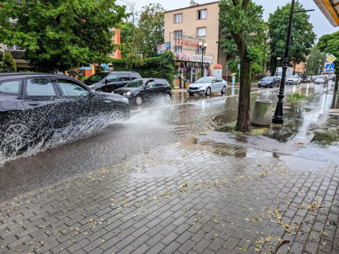 Samochody jadące zalaną ulicą rozpryskują wodę na chodnik