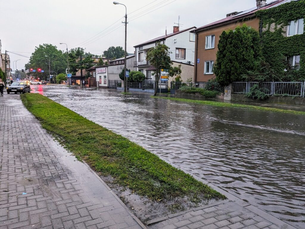 Zalana ulica, którą nie da się przejechać, woda podchodzi pod posesje, w tle sygnalizacja świetlna