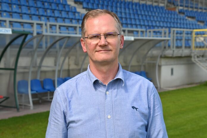 Mężczyzna w średnim wieku w jasnobłękitnej koszuli na tle stadionu piłkarskiego