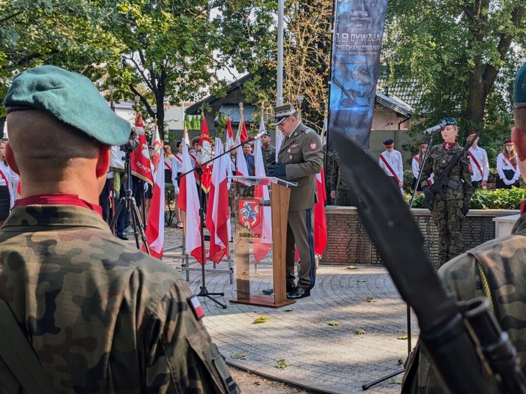 Żołnierz na tle polskich flag przemawia zza mównicy.