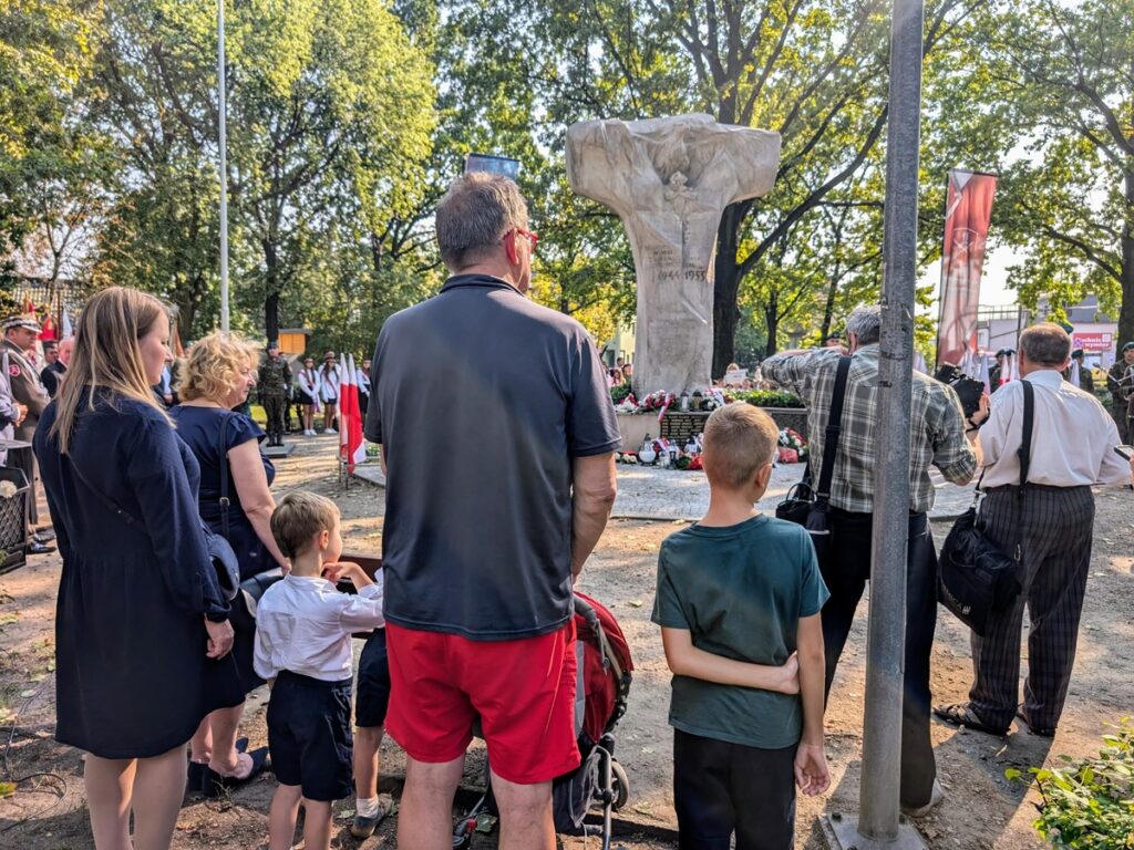 Ludzie w niedużym lasku, stoją w okolicy pomnika kształtem przypominającego krzyż