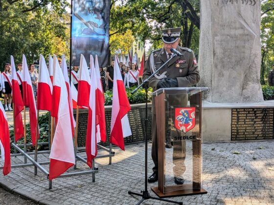 Żołnierz przemawia z mównicy w tle polskich flag w lasku