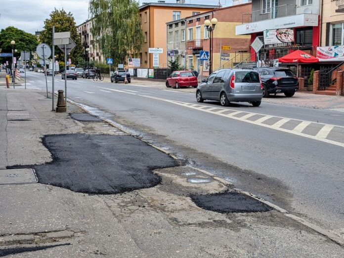 Ulica z dziurą, na której przejeżdżają samochody, wskazująca na potrzebę naprawy nawierzchni.