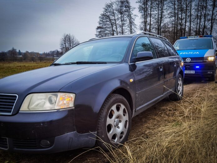 Granatowy samochód osobowy stoi na polnej drodze, za nim auto policyjne