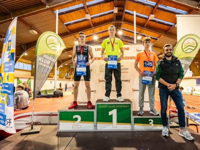 Trzech uśmiechniętych mężczyzn na podium podczas wydarzenia lekkoatletycznego w hali, z medalami.