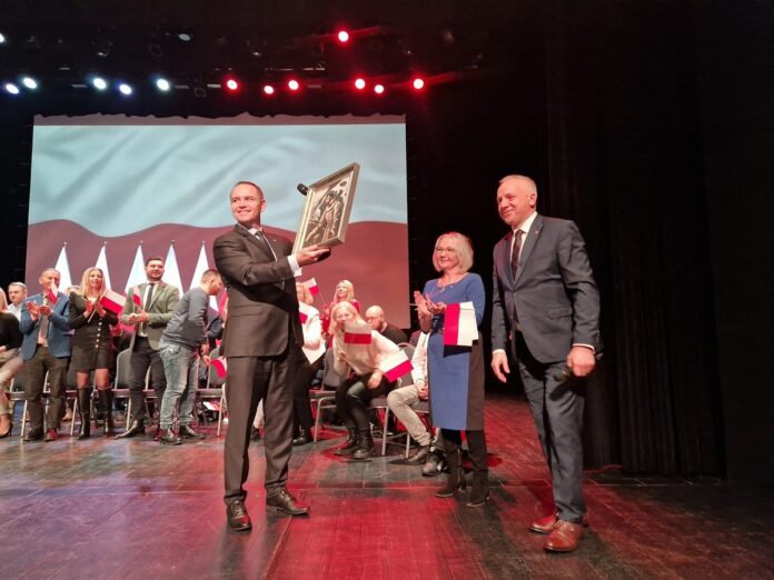 Uśmiechnięty mężczyzna stoi na scenie, w dłoniach trzyma obraz, towarzyszą mu kobieta i mężczyzna, w tle, na telebimie, biało-czerwona flaga