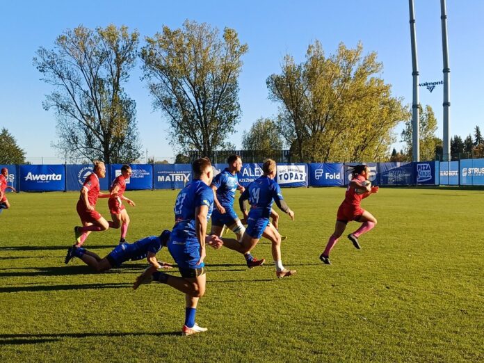 Grupa mężczyzn grających w piłkę rugby na boisku, w dynamicznej akcji sportowej.