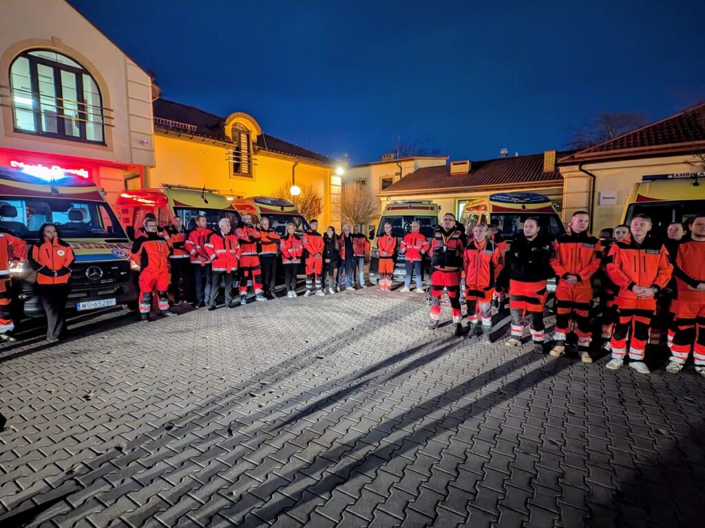 Grupa ratowników medycznych w czerwonych mundurach stoi przed ambulansem, prezentując solidarność.