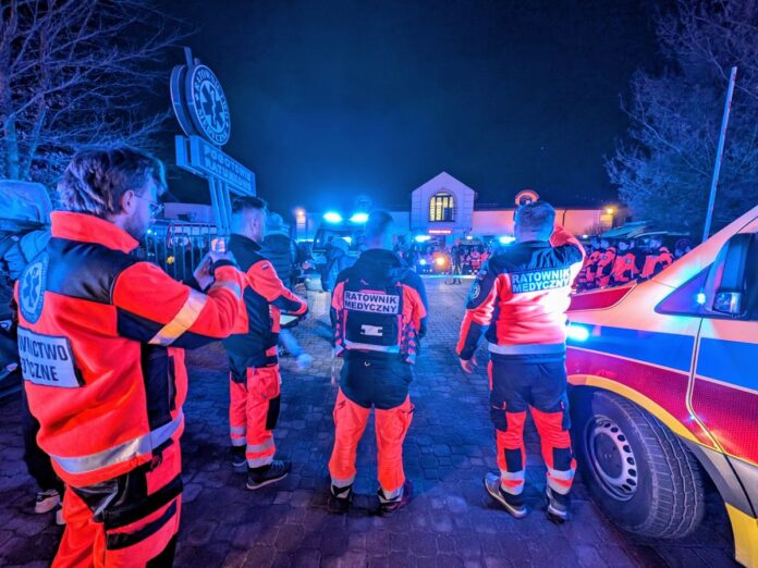 Wieczorową porą grupa ratowników medycznych stoi wokół karetki, obserwując inne ambulanse z włączonymi syrenami.