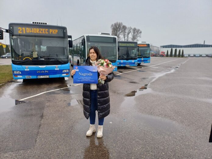 Kobieta stoi przed dwoma autobusami, trzymając bukiet kwiatów w dłoniach,