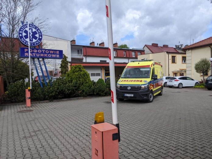 Żółty ambulans wyjeżdżający ze stacji pogotowia, na pierwszym planie podnoszony szlaban.