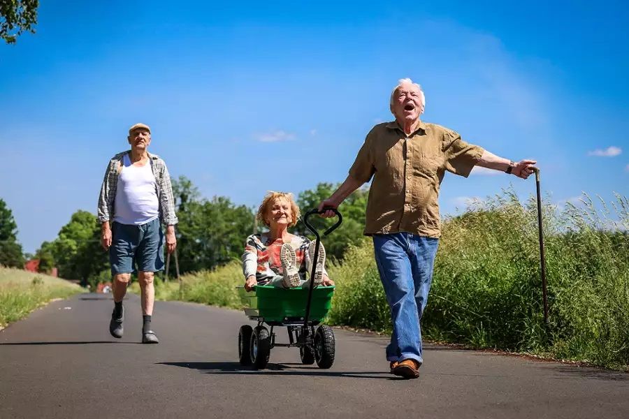 Starszy mężczyzna wiezie kobietę w wózku, ciesząc się wspólnym czasem. Razem z nimi inny mężczyzna. 