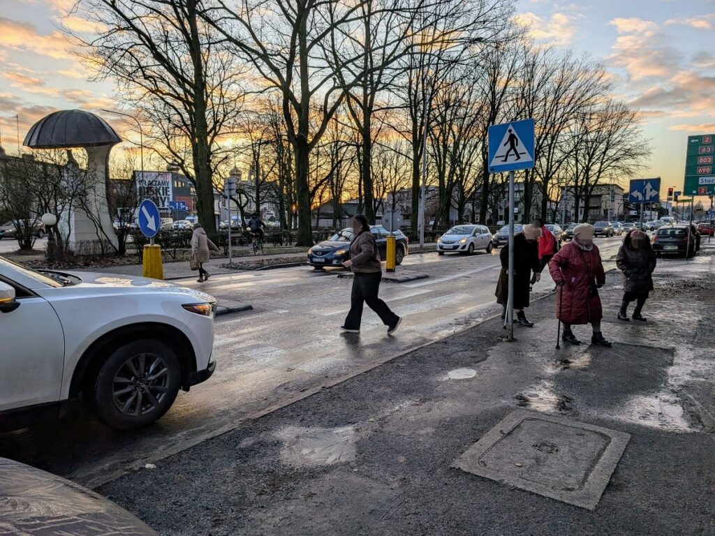 Samochód stojący na ulicy i starsze osoby przechodzące przez jezdnię. 